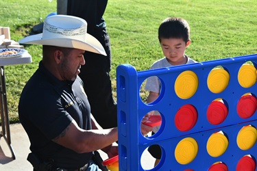 Police officer playing a giant Connect Four game with a child at an outdoor event.