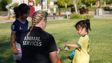 Animal Services officer interacting with a young girl during a community event in a park.