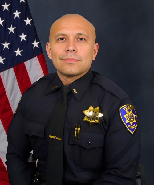 Portrait of a Tracy Police officer in uniform standing in front of an American flag.