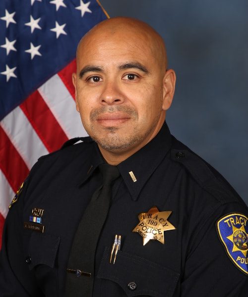 Portrait of a Tracy Police officer in uniform standing in front of an American flag.