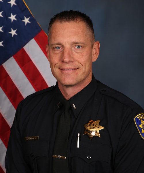 Portrait of a Tracy Police officer in uniform standing in front of an American flag.
