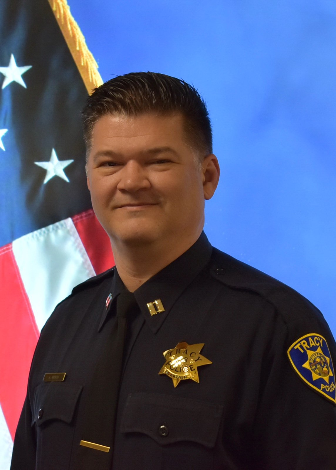 Color portrait of a Tracy police chief in uniform standing in front of American and blue flags.